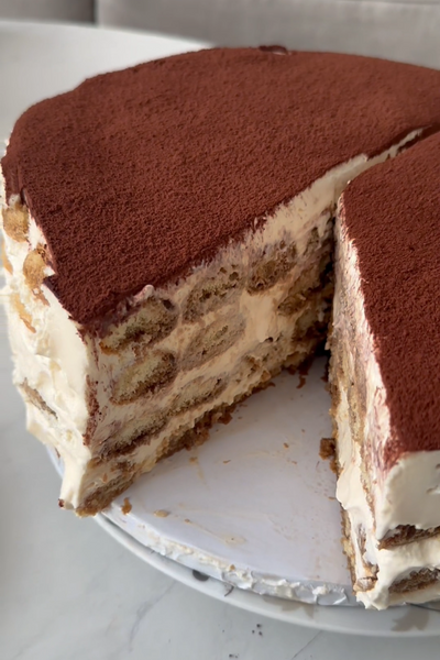 A slice of Tiramisu Cake with layers of sponge and cream, topped with cocoa powder, on a white plate with a piece missing.